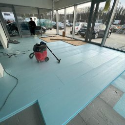 KKBuilding - Układanie niebieskich płyt izolacyjnych pod wylewkę w przestronnym, przeszklonym pomieszczeniu, widoczny odkurzacz przemysłowy i częściowo ułożona podłoga.
