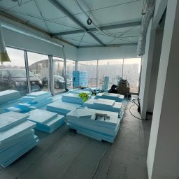 KKBuilding - Przygotowane do montażu płyty styropianowe, ułożone w stosy na podłodze w pomieszczeniu z dużymi oknami, narzędzia na wierzchu jednego ze stosów, widoczne instalacje pod sufitem.
