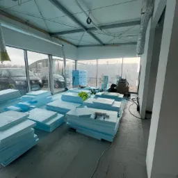 Przygotowane do montażu płyty styropianowe, ułożone w stosy na podłodze w pomieszczeniu z dużymi oknami, narzędzia na wierzchu jednego ze stosów, widoczne instalacje pod sufitem.