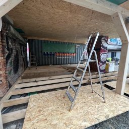 KKBuilding - Wnętrze w trakcie budowy: drewniany szkielet dachu i podłogi z płyt OSB, drabina oparta o ceglany filar z graffiti, kontener z reklamą w tle.