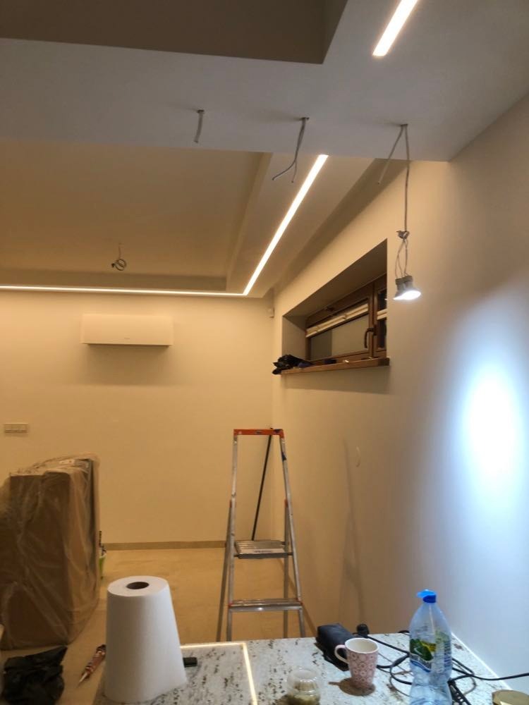 Montaż sufitu z oświetleniem LED w trakcie wykańczania wnętrza, widoczne kable, drabina, zabezpieczona folią meblościanka i akcesoria na blacie.