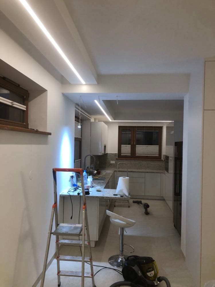 Wnętrze kuchni w trakcie montażu oświetlenia LED podsufitowego, widoczna drabina, narzędzia i odkurzacz, białe ściany i meble.