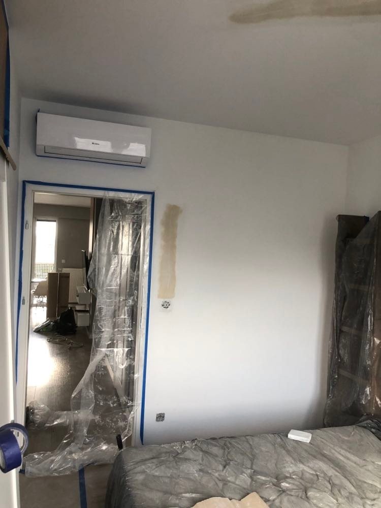 Pokój w trakcie przygotowań do malowania: ściany zabezpieczone taśmą malarską i folią, widoczny klimatyzator, ślady po szpachlowaniu na ścianie.