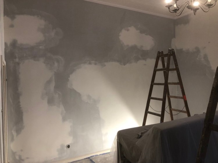 Ściana w trakcie malowania, z widocznymi plamami szarej farby i drewnianą drabiną opartą o ścianę, zabezpieczone meble pod ścianą, widoczny żyrandol na suficie.