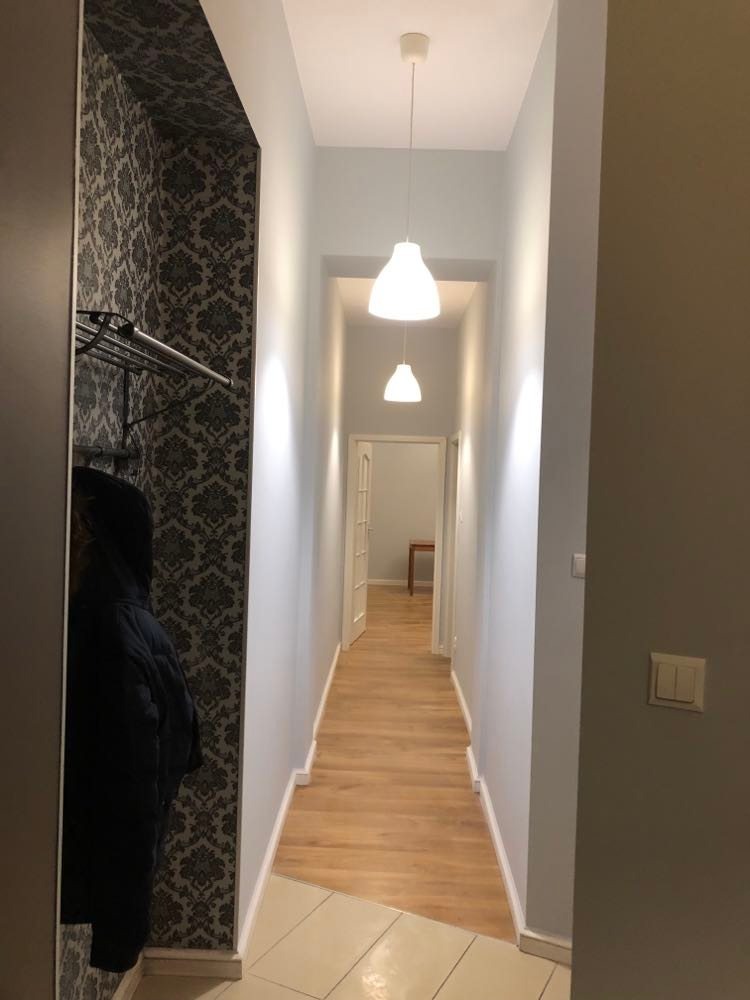 Wąski korytarz po remoncie z drewnianą podłogą, oświetlony dwoma nowoczesnymi lampami wiszącymi, z fragmentem garderoby z wzorzystą tapetą po lewej stronie.