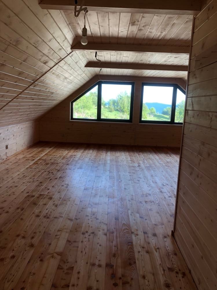 Poddasze wykończone boazerią, z drewnianą podłogą i dwoma trapezoidalnymi oknami z widokiem na zieleń. Widoczna surowa żarówka zwisająca z drewnianego sufitu.
