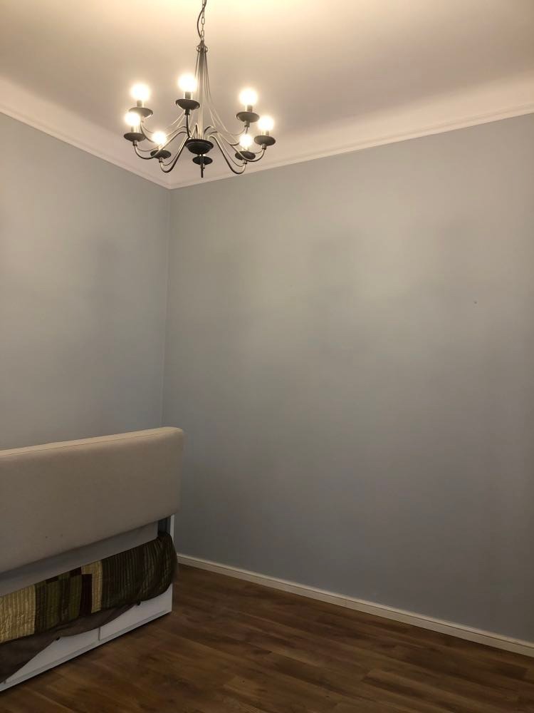 Pomalowane na szaro pomieszczenie z drewnianą podłogą, widoczny fragment sofy i ozdobny żyrandol z żarówkami w stylu świec.