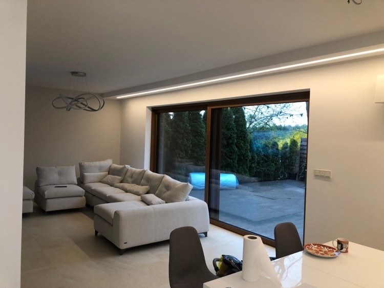 Nowoczesny salon z narożną sofą w odcieniach szarości, oświetleniem LED w suficie i dużym oknem przesuwnym z widokiem na ogród. Na pierwszym planie stół z niedojedzoną pizzą i ręcznikiem papierowym.