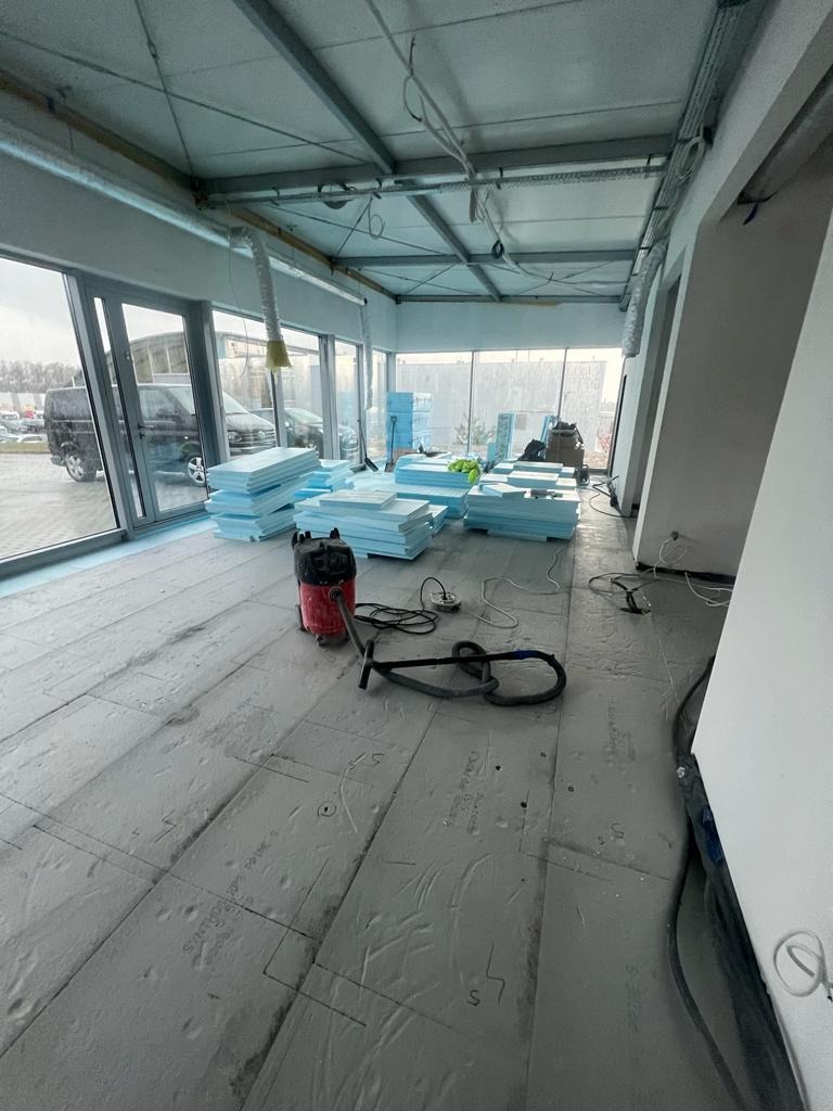 Przygotowanie podłoża w nowym biurze: rozłożone płyty izolacyjne z zaznaczonymi liniami cięcia, odkurzacz przemysłowy i stosy niebieskiego styropianu w przestronnym, jasnym pomieszczeniu z dużymi...