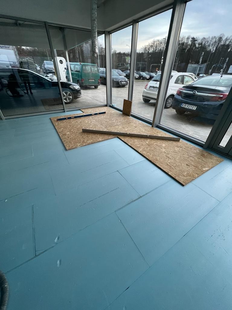 Przygotowanie do montażu podłogi: płyty OSB i poziomice na pomalowanej na niebiesko podłodze w pomieszczeniu z dużymi oknami, widok na parking.