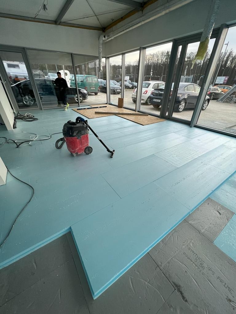 Układanie niebieskich płyt izolacyjnych pod wylewkę w przestronnym, przeszklonym pomieszczeniu, widoczny odkurzacz przemysłowy i częściowo ułożona podłoga.