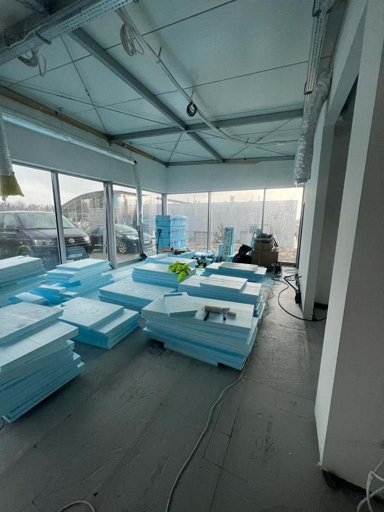 Przygotowane do montażu płyty styropianowe, ułożone w stosy na podłodze w pomieszczeniu z dużymi oknami, narzędzia na wierzchu jednego ze stosów, widoczne instalacje pod sufitem.