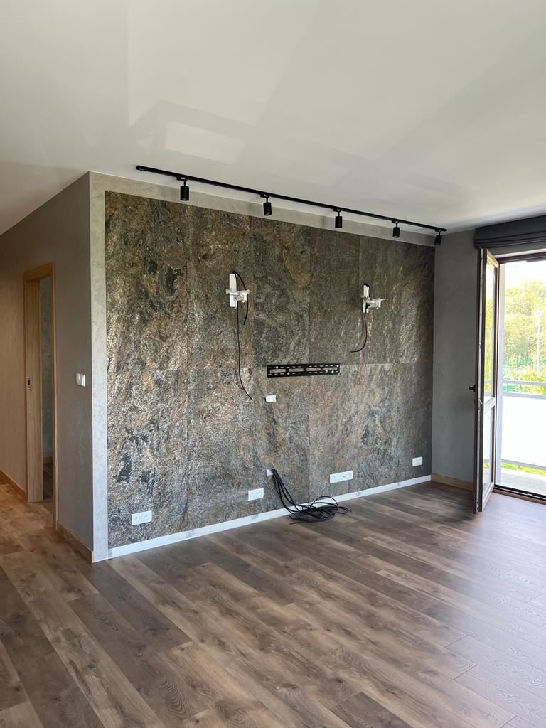 Pokój z wykończoną ścianą imitującą kamień, widoczne gniazdka elektryczne, instalacja pod telewizor i oświetlenie szynowe na suficie.