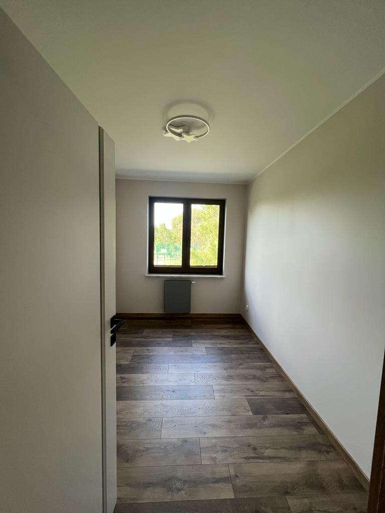 Pusty pokój z panelami podłogowymi imitującymi drewno, pomalowanymi ścianami w odcieniach beżu i białym sufitem z okrągłą lampą. Widoczne okno z widokiem na zieleń i grzejnik pod nim.