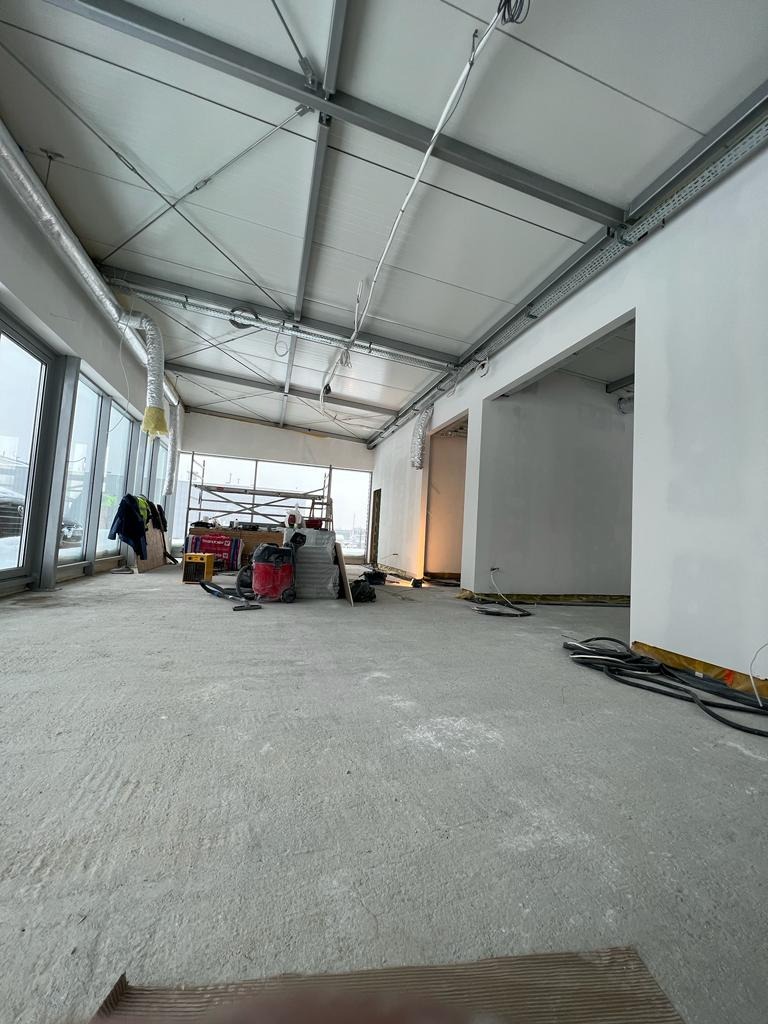 Wnętrze w stanie surowym z widoczną konstrukcją sufitu, przewodami, rusztowaniem w tle oraz narzędziami budowlanymi, ukazujące etap prac wykończeniowych w pomieszczeniu z dużymi oknami.