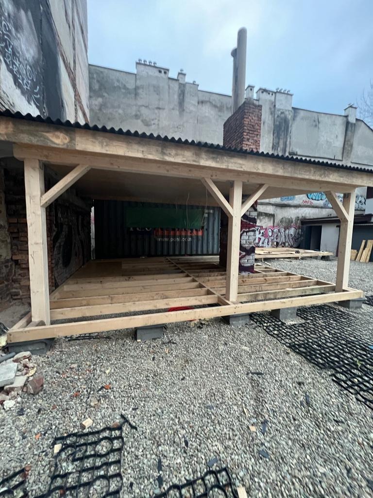 Konstrukcja drewnianego zadaszenia w trakcie budowy, wsparta na betonowych bloczkach, na żwirowym podłożu z elementami geokraty, w tle budynki z graffiti i ceglanym kominem.
