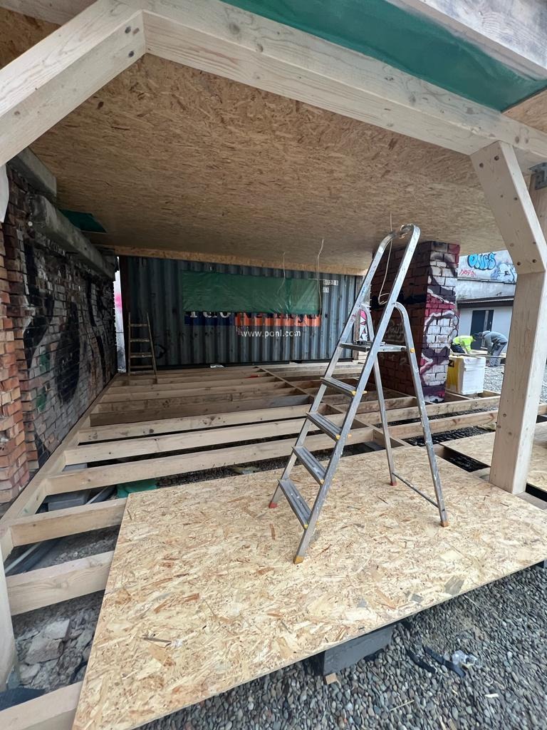 Wnętrze w trakcie budowy: drewniany szkielet dachu i podłogi z płyt OSB, drabina oparta o ceglany filar z graffiti, kontener z reklamą w tle.