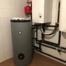 Instalacja grzewcza w kotłowni: szary zbiornik buforowy z termometrem, biały piec gazowy Fujitsu Waterstage, czerwony zbiornik wyrównawczy, czarne rury instalacyjne na tle białych płytek.