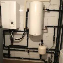 Biała jednostka wewnętrzna pompy ciepła Fujitsu Waterstage z podłączonym zasobnikiem ciepłej wody użytkowej Galmet, rury w izolacji, na tle wykafelkowanej ściany.