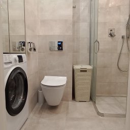 Marcin Oleszczuk - Nowoczesna łazienka z pralką, wiszącą toaletą, kabiną prysznicową i koszem na pranie, wykończona w jasnych, beżowych płytkach imitujących kamień.