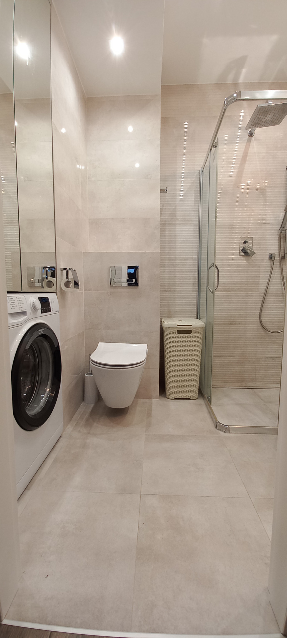 Nowoczesna łazienka z pralką, wiszącą toaletą, kabiną prysznicową i koszem na pranie, wykończona w jasnych, beżowych płytkach imitujących kamień.