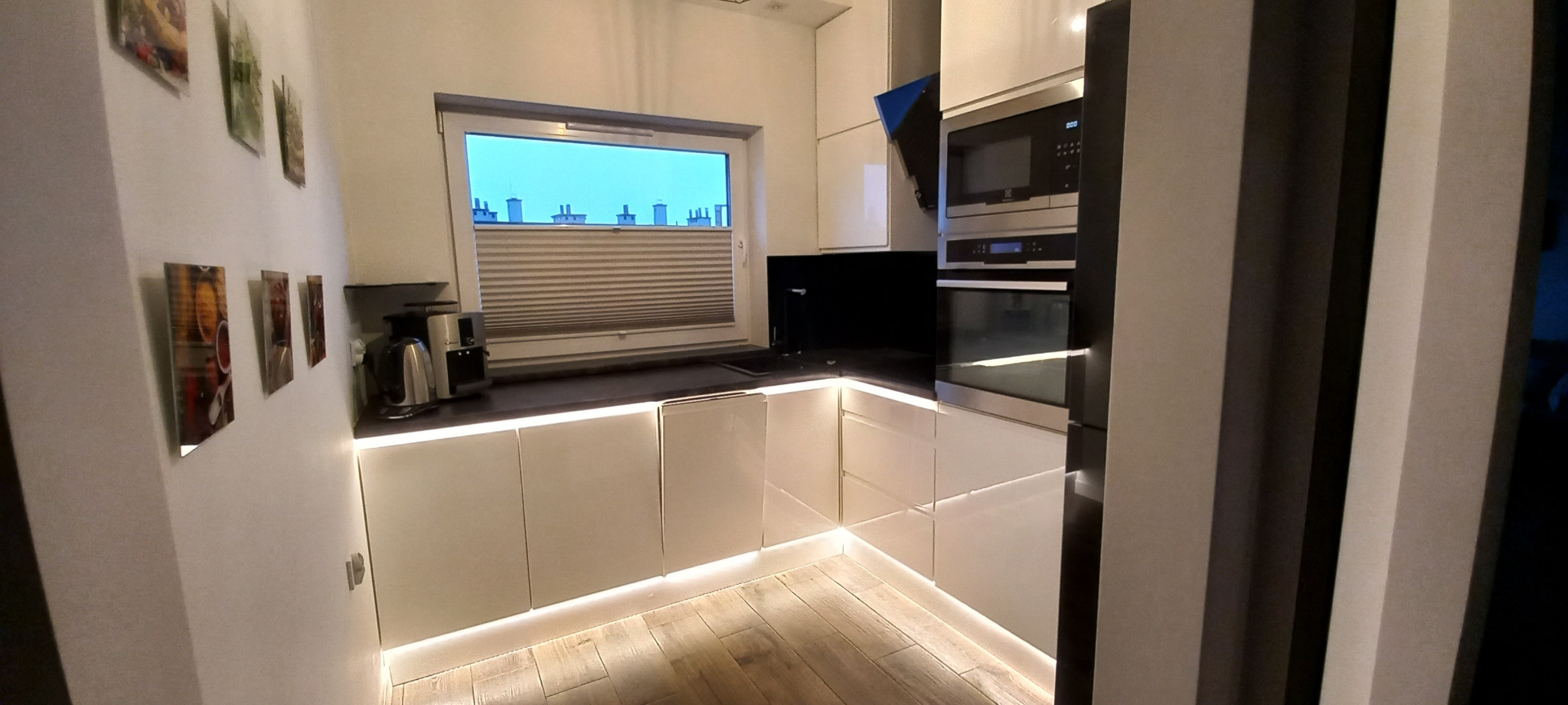 Nowoczesna kuchnia z białymi szafkami podświetlonymi LED, drewnianą podłogą i oknem z widokiem na dachy budynków.