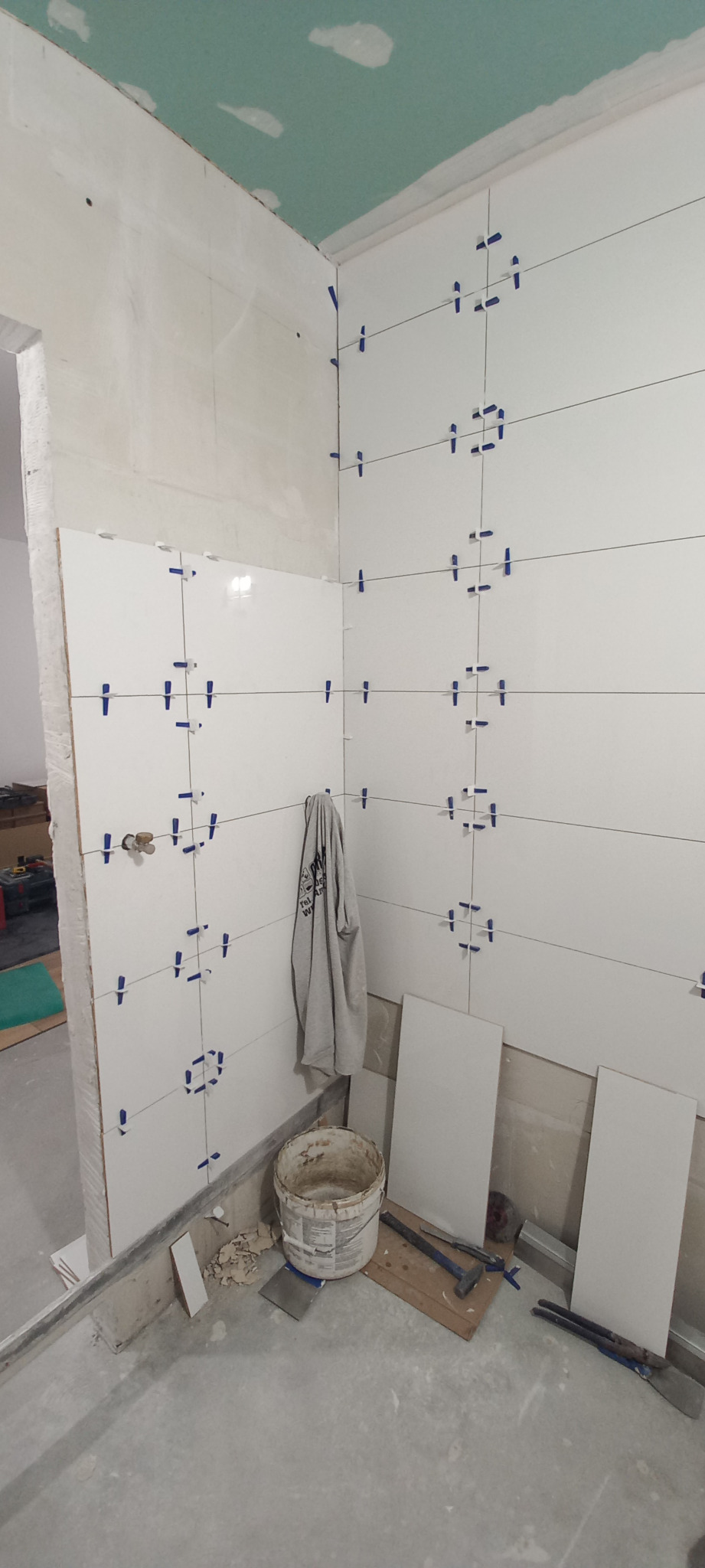 Białe płytki układane z użyciem krzyżyków dystansowych w trakcie prac w łazience w Radzyminie, widoczne narzędzia i wiadro po kleju, sufit częściowo wykończony płytami gipsowo-kartonowymi.