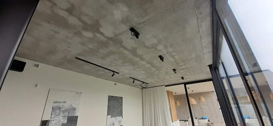 Betonowy sufit z zamontowanym systemem oświetlenia szynowego w nowoczesnym wnętrzu z dużymi oknami.