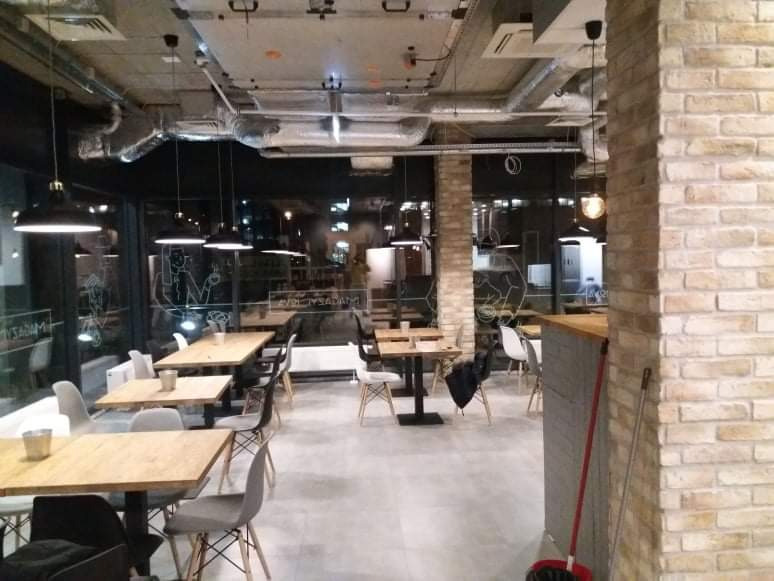 Wnętrze biura z surowym wykończeniem, widoczne drewniane stoły, szare krzesła, ceglane filary i odsłonięte instalacje pod sufitem, duże okna z dekoracyjnymi napisami.