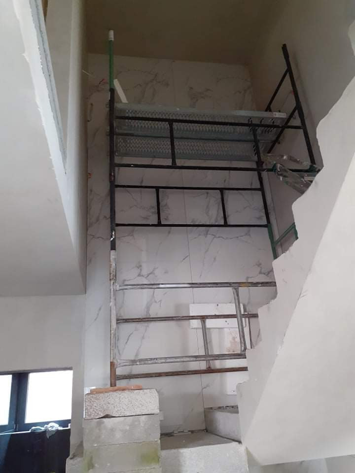 Wysokie rusztowanie ustawione przy ścianie wyłożonej marmurowymi płytkami w trakcie prac wykończeniowych w dwupoziomowym pomieszczeniu ze schodami.