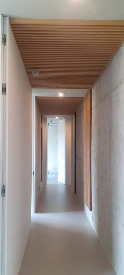 Długi korytarz z betonową ścianą po prawej stronie, drewnianym sufitem lamelowym i otwartymi białymi drzwiami na końcu, oświetlony punktowo.