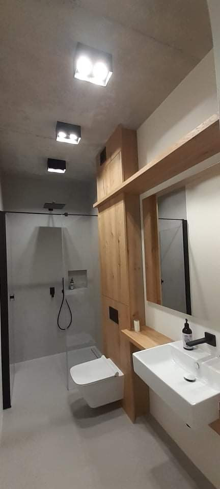 Nowoczesna łazienka z betonową fakturą sufitu, kabiną prysznicową typu walk-in z czarnymi detalami, podwieszaną toaletą i umywalką nablatową na drewnianym blacie.