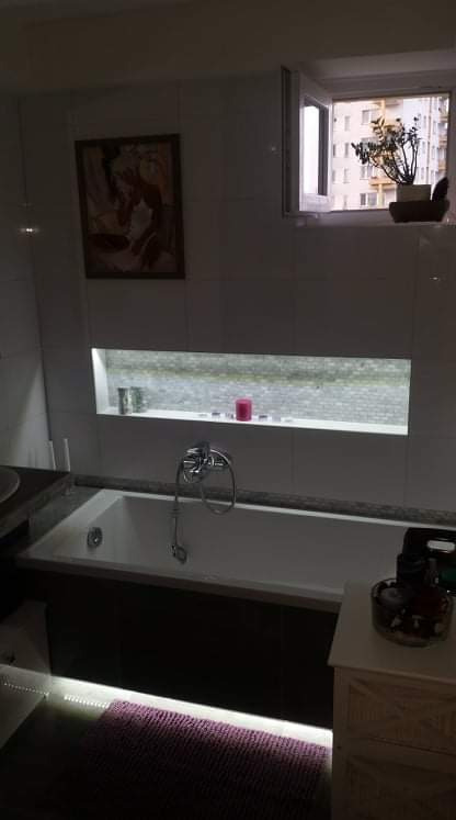 Nowoczesna łazienka z podświetlaną wanną, dekoracyjną wnęką nad wanną z akcentami i obrazem na ścianie, widok na budynek przez okno z rośliną w doniczce.