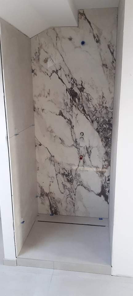 Świeżo wykafelkowana kabina prysznicowa z imitacją marmuru na ścianach, widoczne otwory na armaturę i delikatne niebieskie zabezpieczenia na krawędziach płytek.