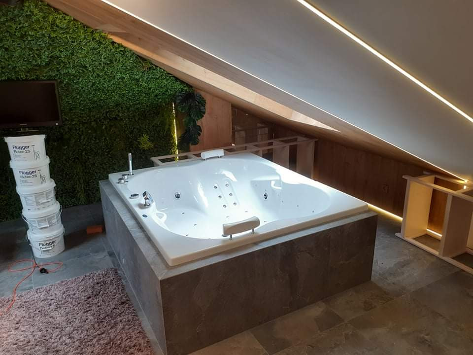 Wanna z hydromasażem zabudowana szarą płytą w łazience na poddaszu z oświetleniem LED w suficie i zieloną ścianą z roślin, obok ustawione wiaderka z farbą