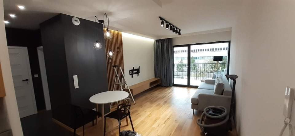 Jasne wnętrze mieszkania po remoncie z parkietem, czarną ścianą działową, drewnianymi lamelami, oświetleniem punktowym i wiszącymi lampami, częściowo umeblowane, z drabiną opartą o ścianę.