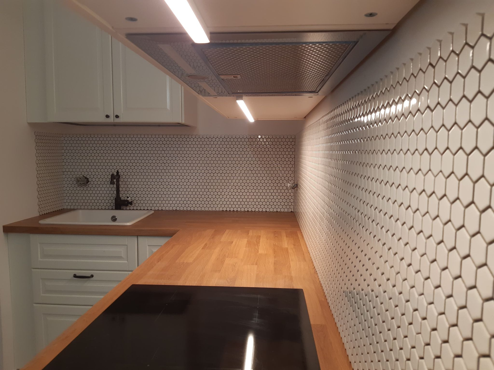 Wykończona kuchnia z białymi szafkami, drewnianym blatem i heksagonalnymi płytkami na ścianie, widok na zlew z czarną baterią i czarną płytą indukcyjną.