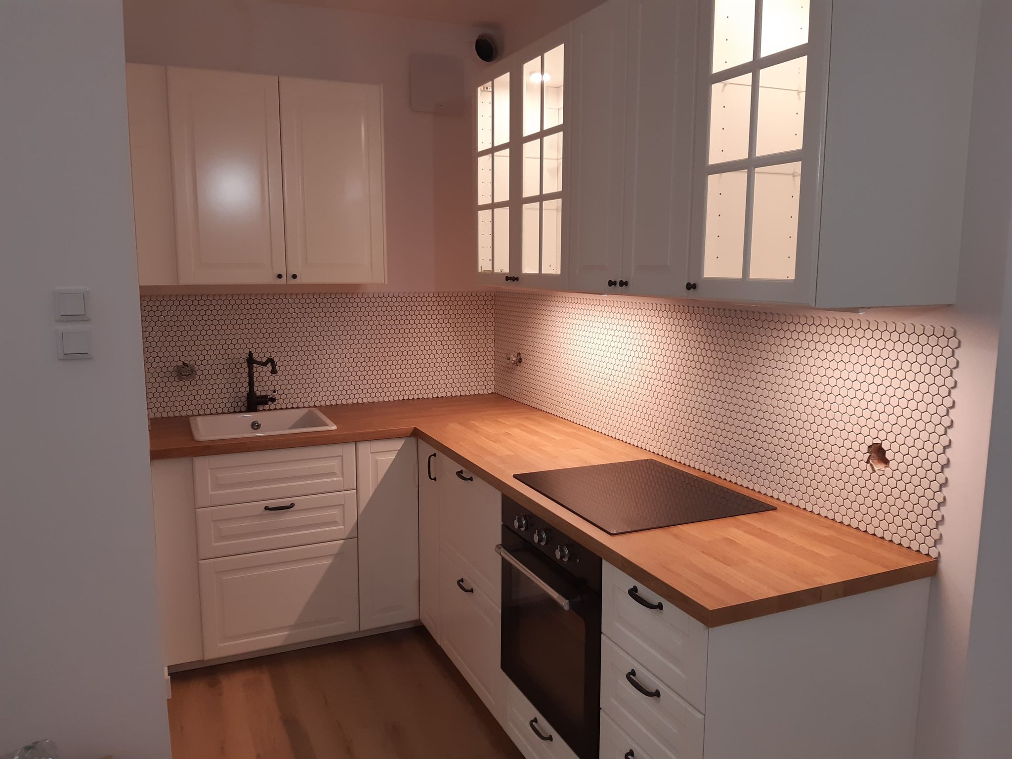 Nowoczesna kuchnia z białymi szafkami, drewnianym blatem i mozaiką heksagonalną na ścianie, czarna armatura i płyta indukcyjna.