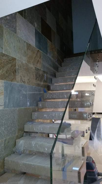 Wykończone schody z kamienną okładziną ściany, zabezpieczone folią ochronną i szklaną balustradą, widoczny odkurzacz na podłodze.