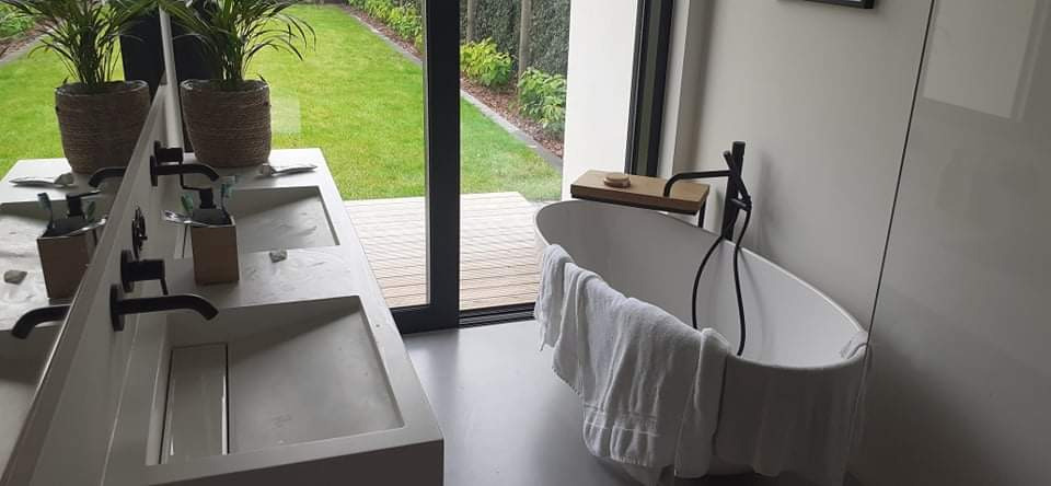 Nowoczesna łazienka z podwójną umywalką z betonu architektonicznego i wolnostojącą wanną na tle okna z widokiem na ogród.