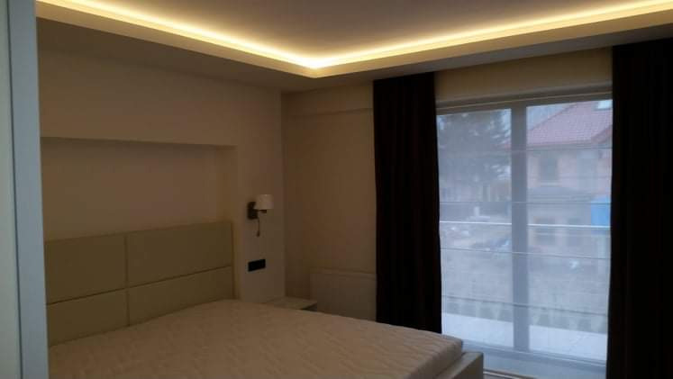 Nowoczesna sypialnia z podświetlanym sufitem LED, beżowe panele zagłówka łóżka, ciemne zasłony i widok na budynek przez okno z roletami rzymskimi.