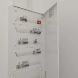 Otwarta, biała skrzynka elektryczna z widocznymi bezpiecznikami i wyłącznikami, zamontowana na białej ścianie, z widocznym kablem uziemiającym.