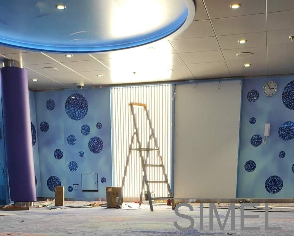 Wnętrze pomieszczenia w trakcie prac wykończeniowych: drabina stoi przy oknie z żaluzjami, widoczna kamera monitoringu pod sufitem, ściany z motywem błękitnych kół, zegar i domofon na ścianie...