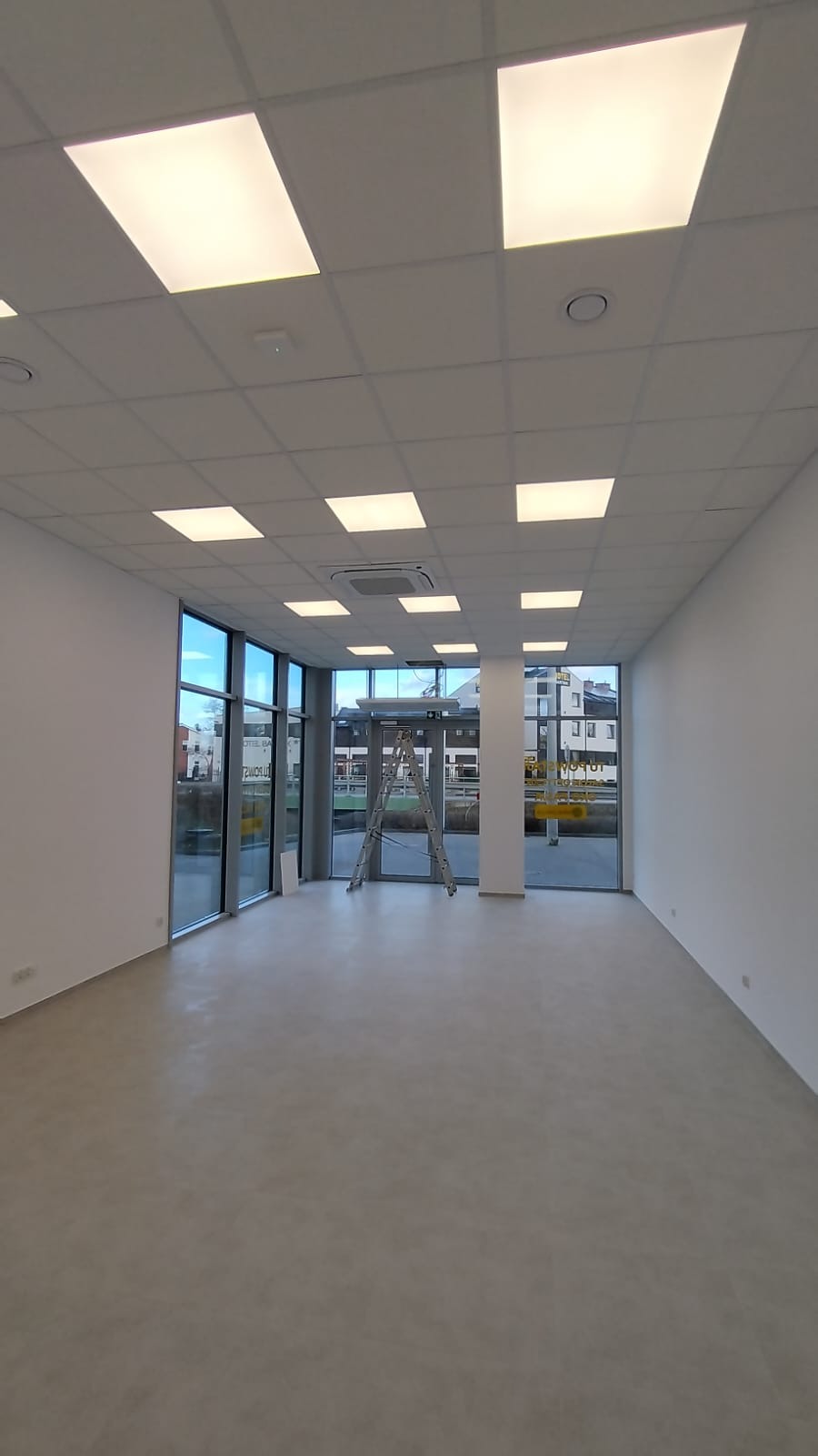 Wnętrze pustego pomieszczenia z zamontowanym systemem oświetlenia rastrowego w suficie podwieszanym, drabina aluminiowa ustawiona przy przeszklonym wejściu.