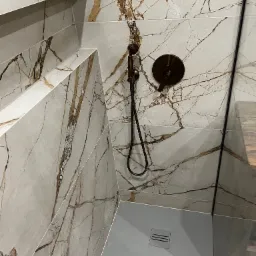 Kabina prysznicowa wykończona płytkami imitującymi marmur z brązowymi żyłami, wyposażona w deszczownicę w kolorze starego złota i odpływ liniowy.