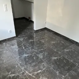 Błyszcząca, ciemna podłoga z płytek imitujących marmur, ułożona w nowym, pustym pomieszczeniu z białymi ścianami i widocznym wejściem do garderoby z półkami.