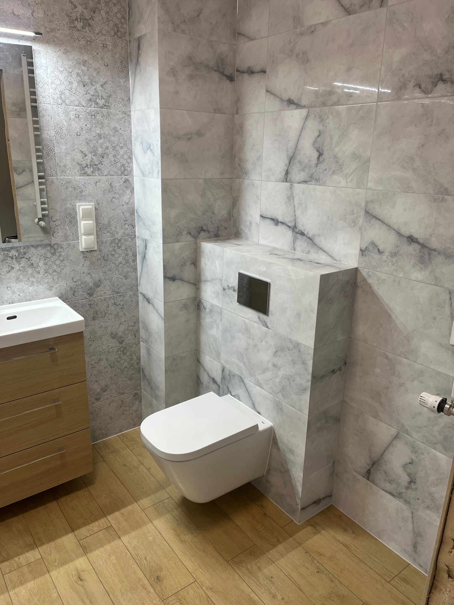 Nowoczesna łazienka z podwieszaną toaletą, marmurowe płytki na ścianach, drewnopodobna podłoga, szafka pod umywalką z drewnianymi frontami, minimalistyczny design.