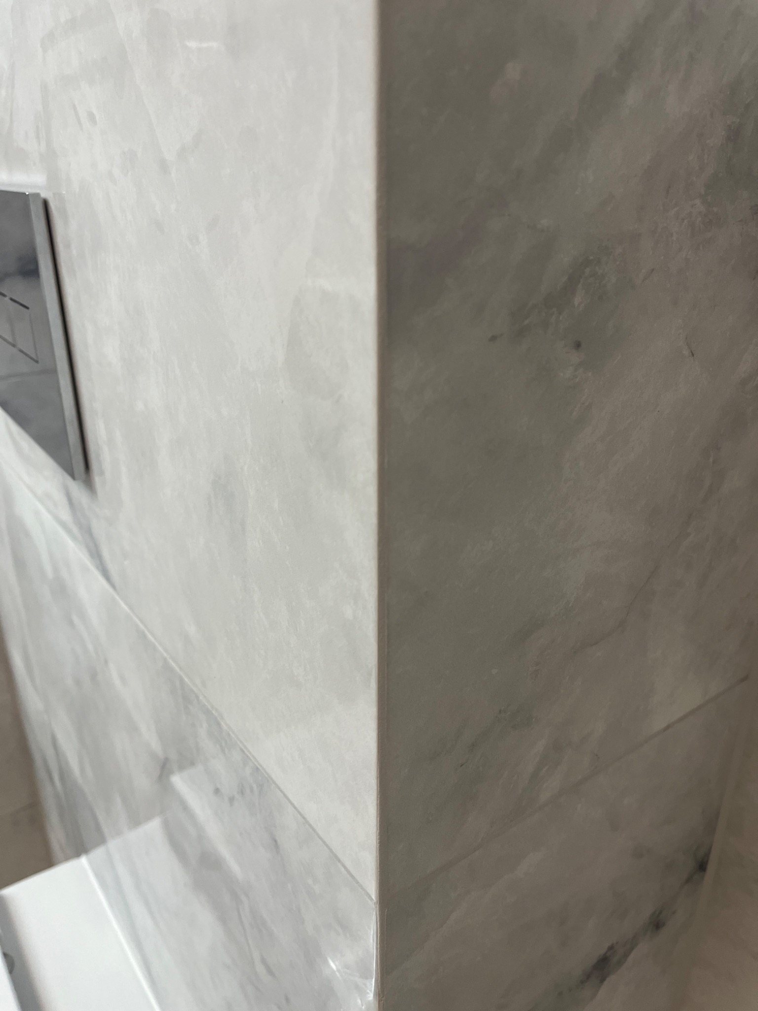 Fragment ściany łazienki wykończonej jasnymi, marmurkowymi płytkami ceramicznymi z widocznym przyciskiem spłuczki.