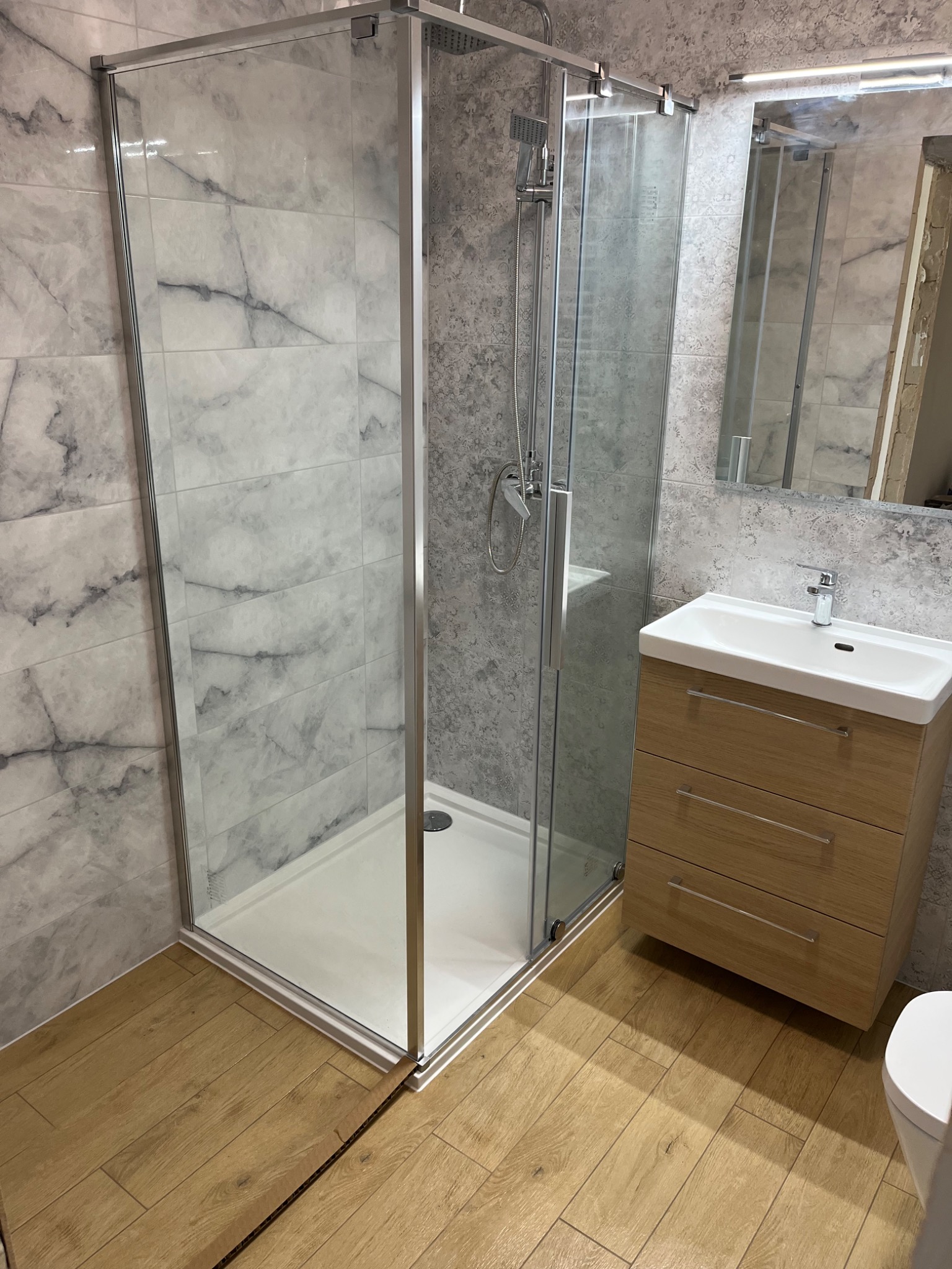 Nowoczesna łazienka po remoncie: kabina prysznicowa z transparentnego szkła, umywalka z drewnianą szafką, marmurowe płytki na ścianach i drewnopodobna podłoga.