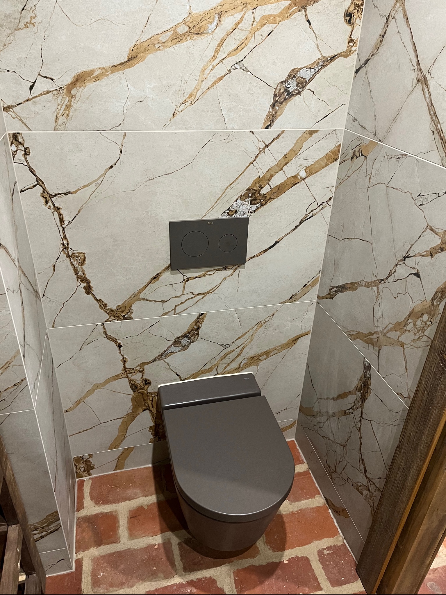 Szara wisząca toaleta z podtynkowym systemem spłukiwania na tle ściany wyłożonej płytkami imitującymi marmur z brązowymi żyłami, podłoga wyłożona czerwoną cegłą.